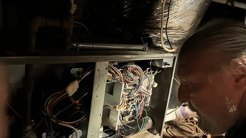 Man inspecting an open furnace with many colorful wires and an exposed circuit board, in a dimly lit basement.